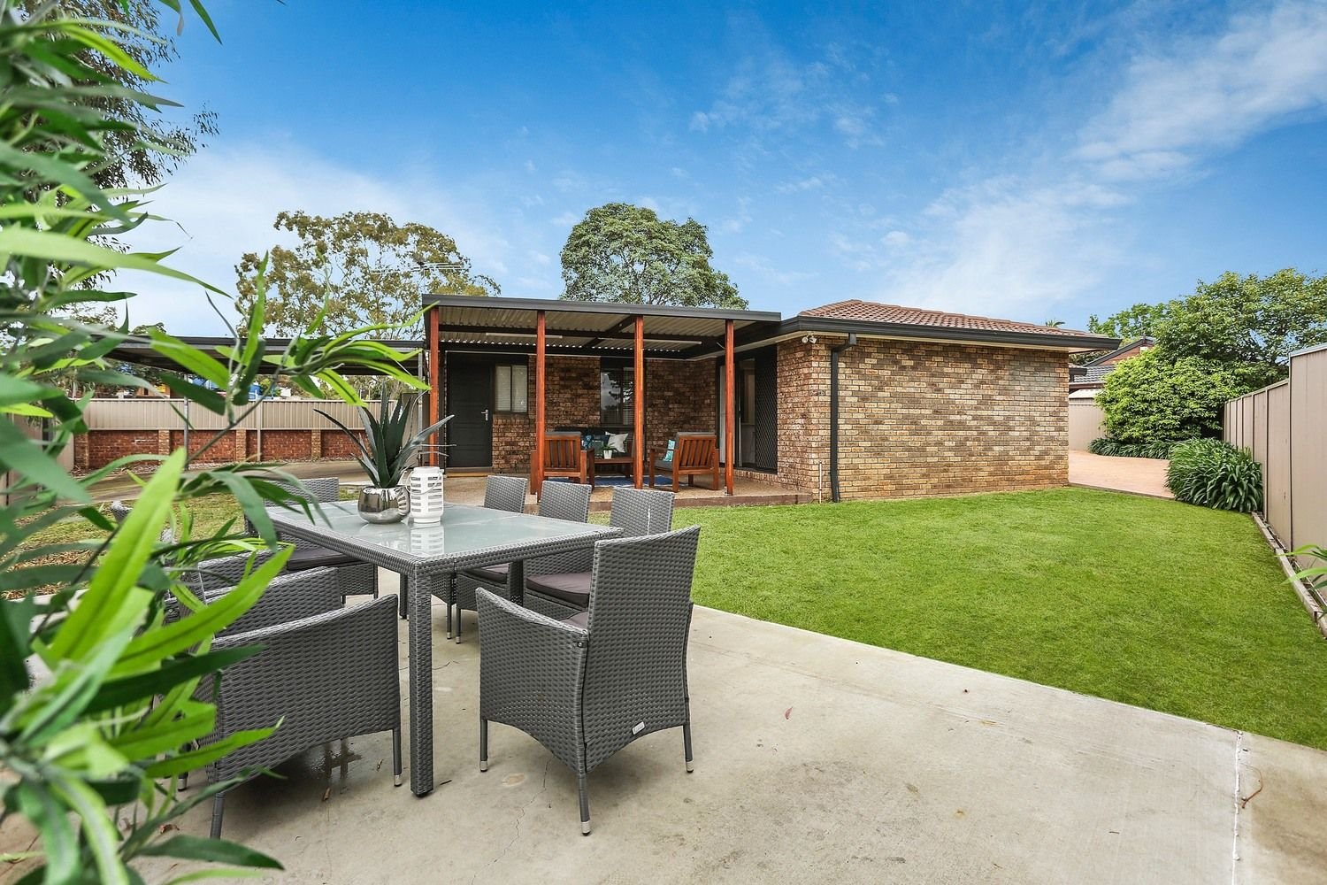 Backyard patio with outdoor dining table, chairs, and a covered seating area with wooden beams, grass lawn, and a brick house with a tiled roof under a blue sky.