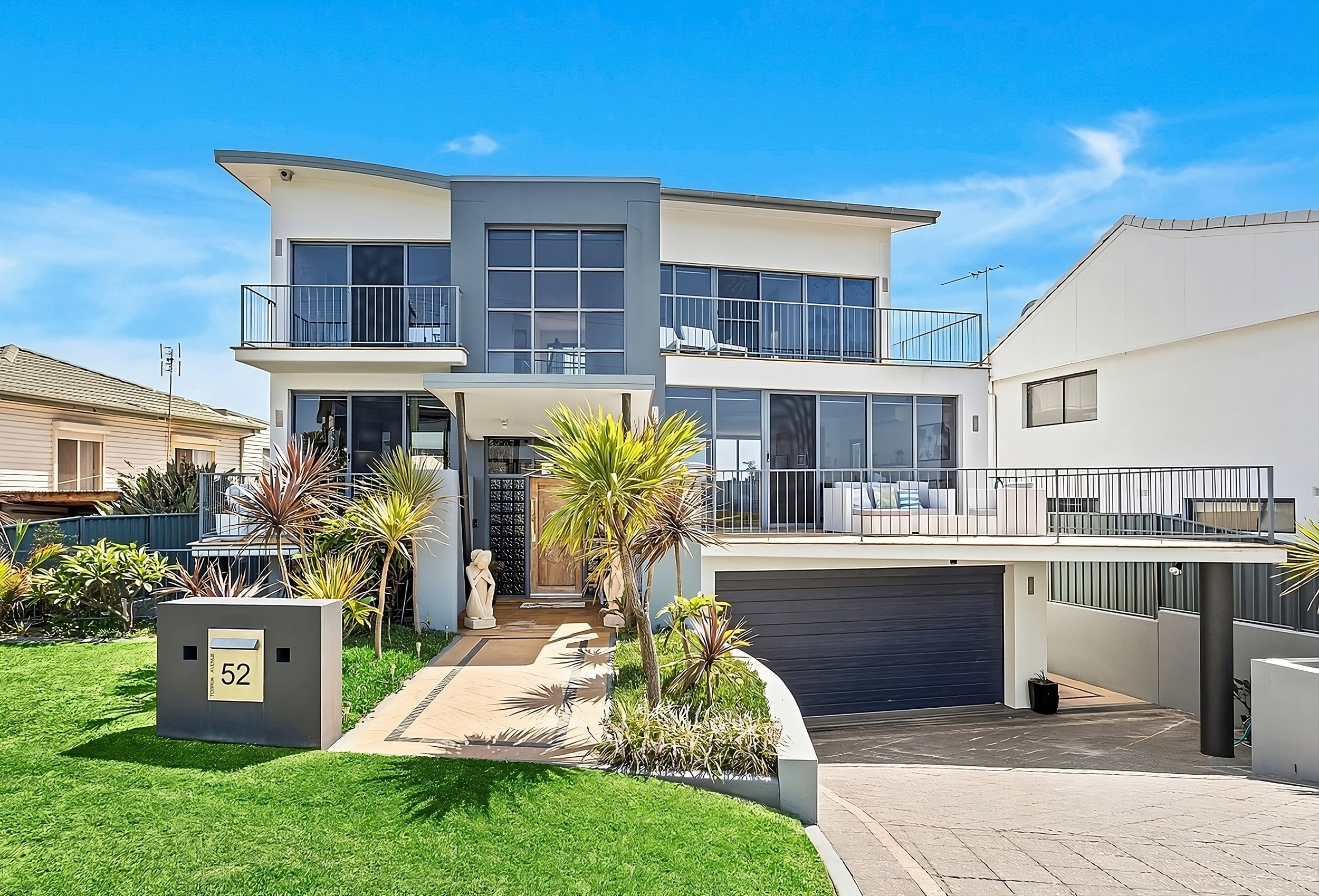Modern multi-story house with large glass windows, balconies, and a garage, surrounded by a well-maintained lawn and tropical plants.