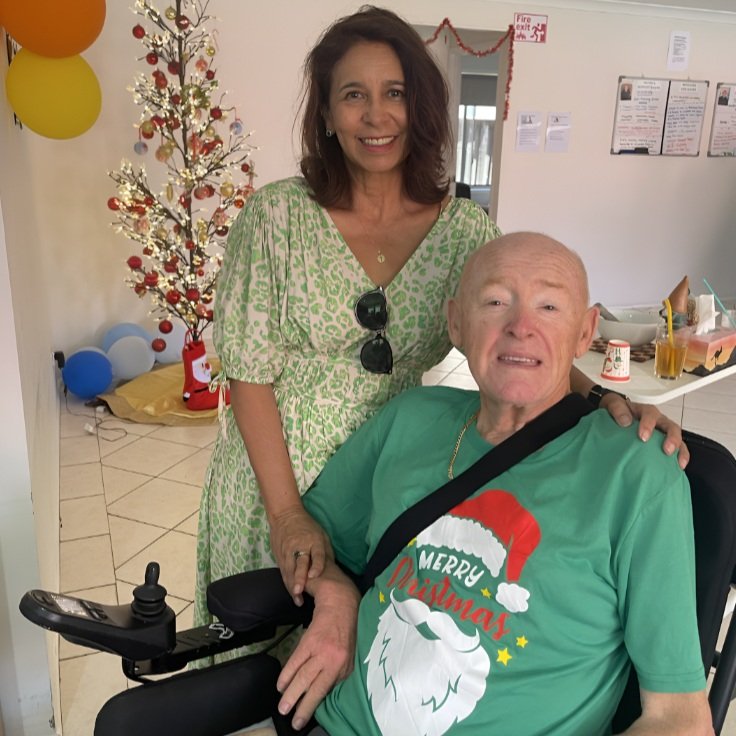 A woman and an elderly man smiling at a Christmas celebration. The woman stands behind the man, who is seated in a wheelchair, and has her hand on his arm. The man wears a green Christmas shirt with a Santa graphic and text that says 'Merry Christmas'. The woman wears a light green dress and sunglasses hanging from her neckline. In the background, there is a decorated Christmas tree, balloons, and holiday decorations.