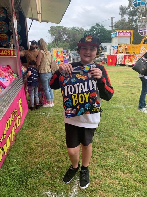 A boy at a fair holding a colorful toy bag and smiling. In the background, there are people shopping and amusement rides.